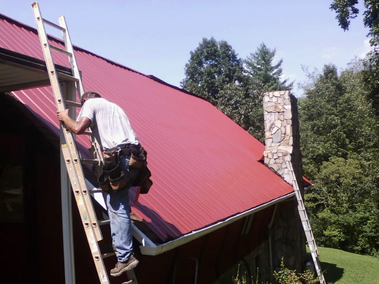 Roofer on a ladder beside a red metal roof and stone chimney — Soffit & Fascia Repair by Roofing Brothers in Arroyo Hondo
