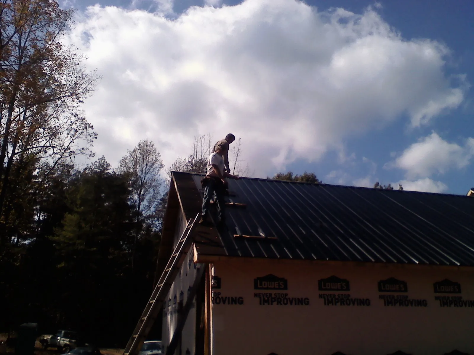 Roofer fastening dark metal panels high against a cloudy sky, from a Roof Inspection job in Arroyo Hondo